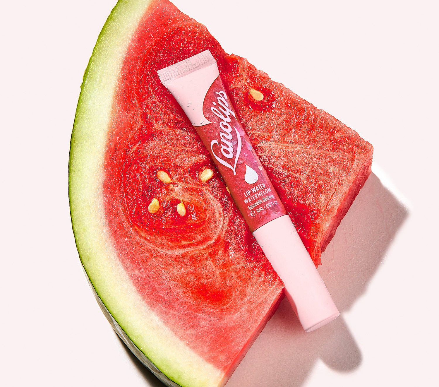 A tube of Lanolips Lip Water Watermelon rests on a slice of fresh watermelon, its vibrant pink packaging complimenting the fruit's rich red tones. This lightweight, hydrating lip serum delivers a transparent watermelon-pink holographic shimmer, giving lips a juicy, glossy glow. Made with liquid lanolin and hyaluronic acid, it boosts moisture while providing a sheer, fruity tint. Like watermelon juice for your lips, this non-sticky, natural formula keeps them soft and hydrated.