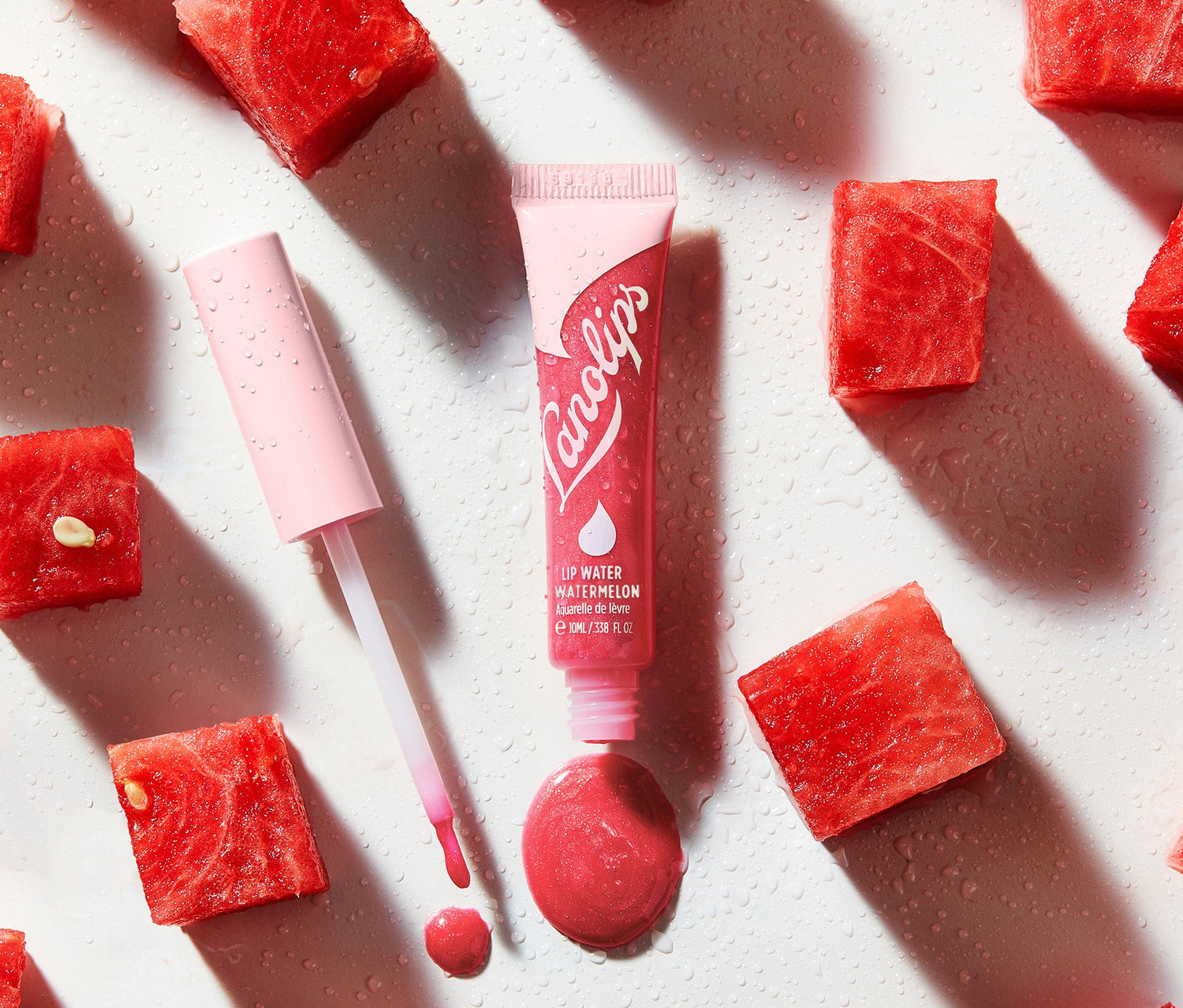 A tube of Lanolips LIp Water Watermelon is placed on a dewy white surface, surrounded by fresh watermelon cubes. The pink tube, with bold light pink Lanolips branding, is uncapped, revealing the applicator with a swipe of the hydrating, glossy formula. This lightweight lip serum delivers a transparent watermelon-pink holographic shimmer, giving lips a juicy, moisturised glow. Made with liquid lanolin and hyaluronic acid, it deeply hydrates while the sheer, fruity tint enhances lips naturally.