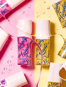 Lifestyle image of Lanolips Hyaluronic Lip Oils in Honey and Raspberry shades, arranged on a pink and yellow split background with oil droplets and flowing lip oil creating a dynamic composition