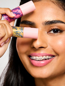 Smiling model holding two Lanolips Hyaluronic Lip Oil shades, highlighting the product's sleek pink packaging. The lip oils, infused with lanolin, hyaluronic acid, and vitamin E, deliver a glossy and nourishing finish for soft, hydrated lips