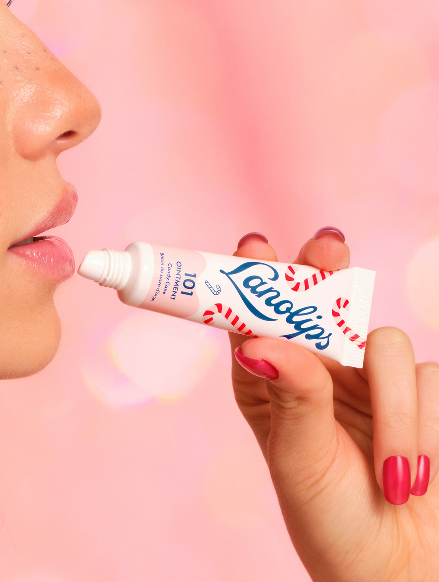 Model applying Lanolips 101 Ointment Multi-Balm Candy Cane lip balm to their lips with a pink gradient background. Hydrates dry lips, skin patches, cuticles, elbows & more. An all-natural lanolin balm with vitamin-e.