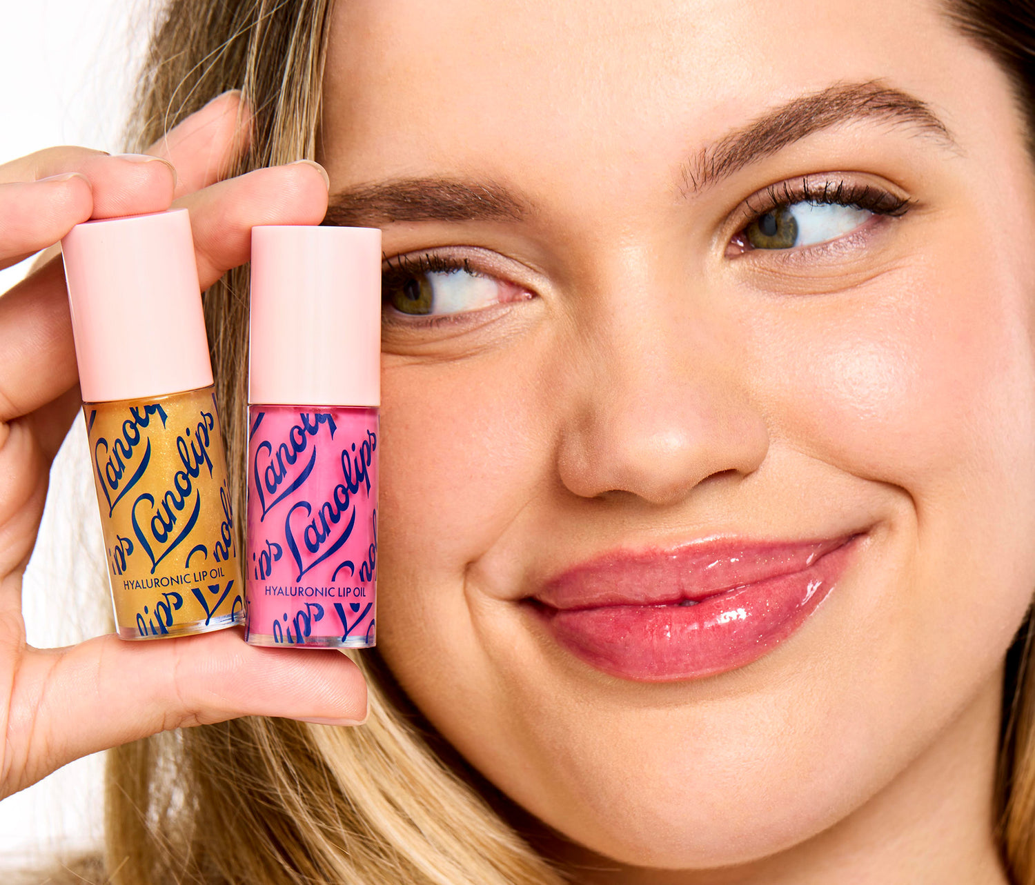 Model holding the tubes Lanolips Hyaluronic Lip Oil Duo in raspberry and honey against her face. The lip oil contains hydrating lanolin, vitamin e and hyaluronic oil for minimum stickiness, and maximum juiciness.