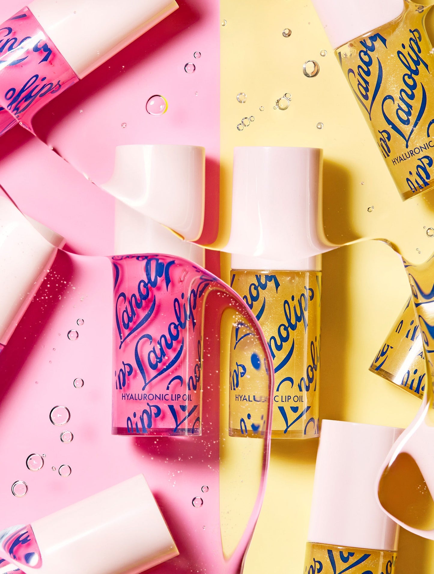 Lifestyle image of Lanolips Hyaluronic Lip Oils in Honey and Raspberry shades, arranged on a pink and yellow split background with oil droplets and flowing lip oil creating a dynamic composition