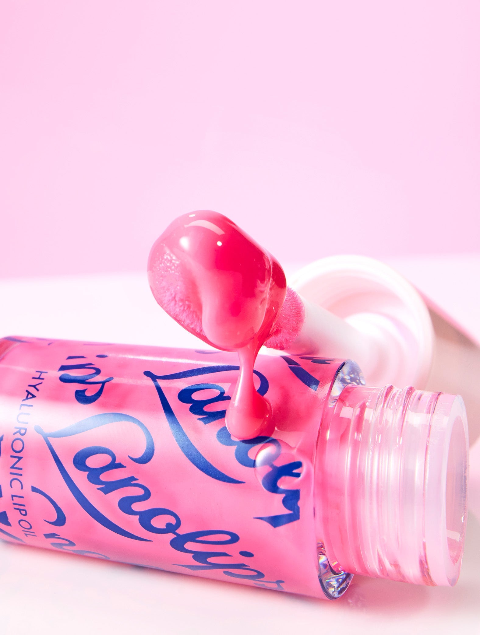 Lifestyle image of Lanolips Hyaluronic Lip Oil in the shade Raspberry, arranged on a yellow background showing the lip oil droplets and lip oil flowing from the container for a dynamic, glossy effect.