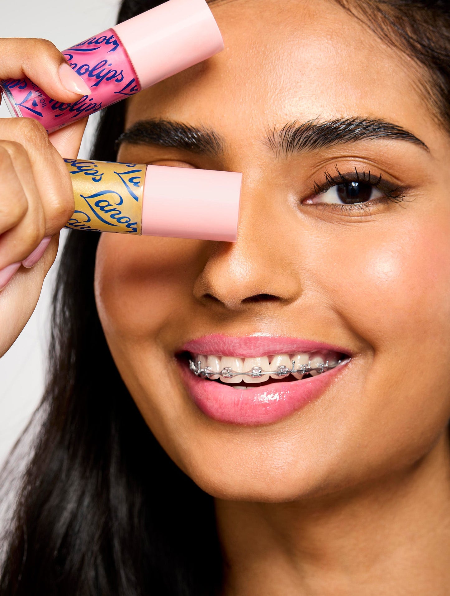 Smiling model holding two Lanolips Hyaluronic Lip Oil shades Raspberry and Honey, highlighting the product's sleek pink packaging. The lip oils, infused with lanolin, hyaluronic acid, and vitamin E, deliver a glossy and nourishing finish for soft, hydrated lips