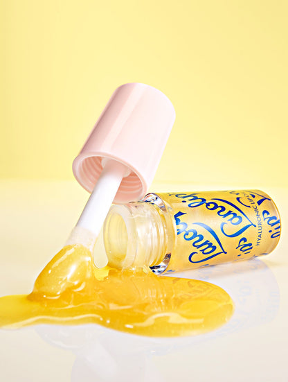 Lifestyle image of Lanolips Hyaluronic Lip Oil in the shade Honey, arranged on a yellow background showing the lip oil droplets and lip oil flowing from the container for a dynamic, glossy effect.