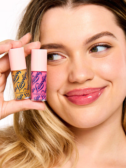 Smiling model holding two Lanolips Hyaluronic Lip Oil shades, highlighting the product's sleek pink packaging. The lip oils, infused with lanolin, hyaluronic acid, and vitamin E, deliver a glossy and nourishing finish for soft, hydrated lips